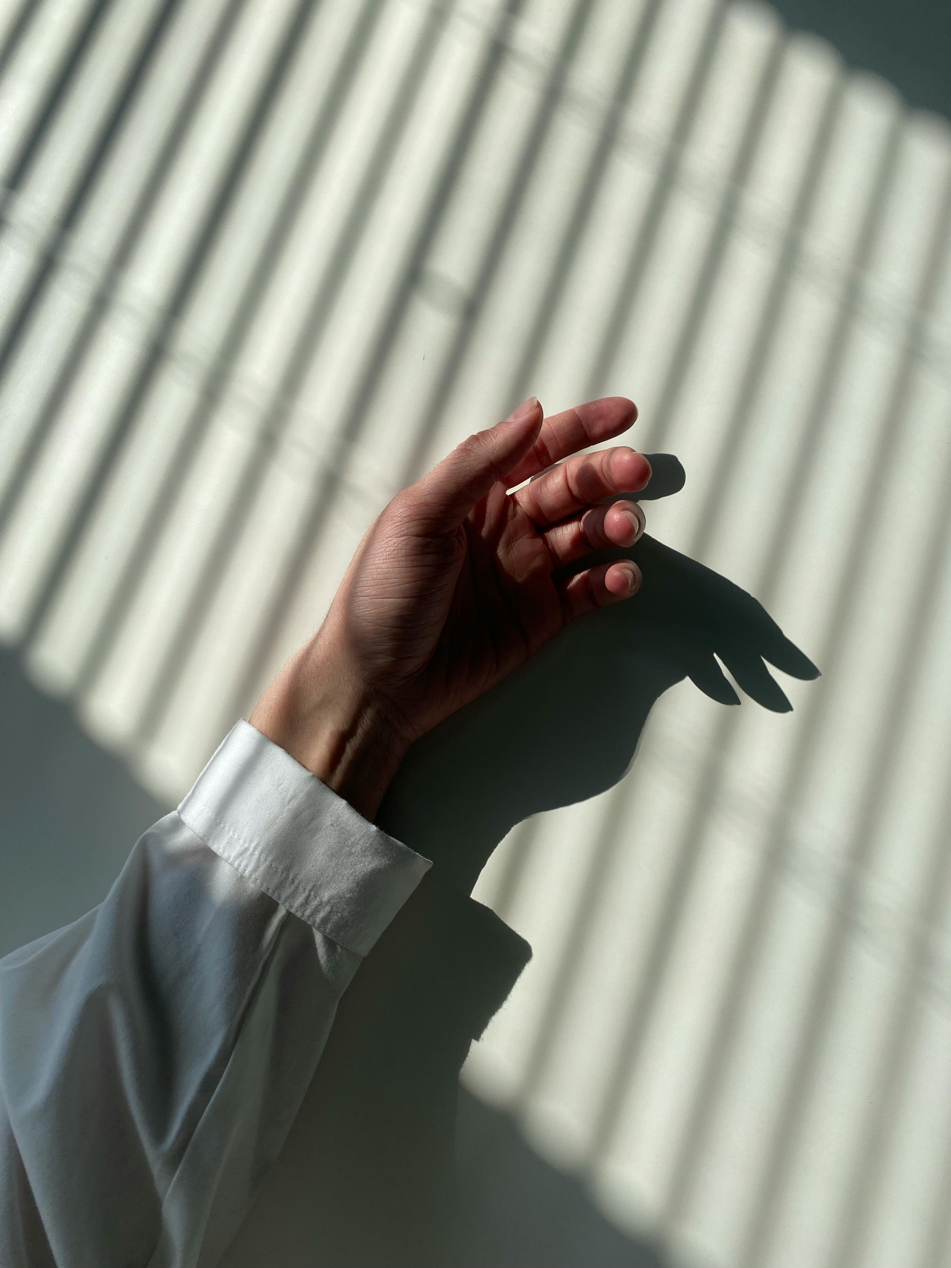 A hand at rest, lit by hard window light from the right; layered architectural shadows fall across the wall.